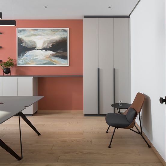 Ein Büroraum mit roter Wand, grauem Schreibtisch und braunem Stuhl. Ein Gemälde hängt über dem Schreibtisch, und eine Topfpflanze steht auf der Theke.