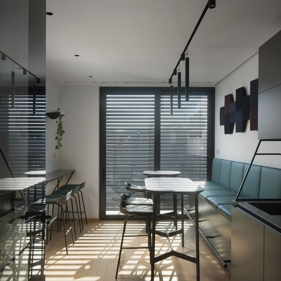 Ein Innenbereich für Mahlzeiten mit weißen Wänden und Holzböden. Es gibt Tische und Stühle, eine Bank und eine Theke. Die Decke hat Leuchten und die Wand Kunstwerke. Es gibt Glastüren mit Jalousien.