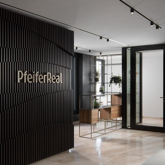Ein Innenraum eines modernen Büroraums mit einer großen schwarzen Wand, auf der das Wort PfeiferReal steht. Der Raum wird von Deckenleuchten beleuchtet und verfügt über einen hölzernen Schreibtisch mit Pflanzen.