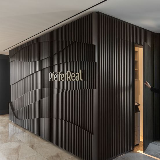 Ein modernes Innendesign zeigt eine große, wellenförmige, dunkelbraune Wand mit dem Logo 'PfeiferReal'. Eine offene Tür offenbart einen Raum mit einem Schrank, und eine Person steht im Flur.