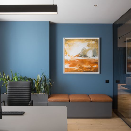 Ein Büroraum mit blauen Wänden verfügt über einen Schreibtisch, einen schwarzen Bürostuhl und eine Lederbank. Ein gerahmtes Gemälde schmückt die Wand, ergänzt durch Topfpflanzen.