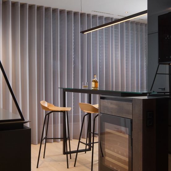 Ein moderner Barbereich mit einem Glastisch, zwei Stühlen, einer Flasche und einem Weinkühler. Dahinter befindet sich ein langer grauer Vorhang.