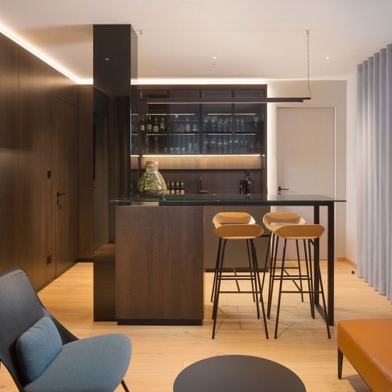 Eine moderne Küche mit Holzboden, einer Bartheke, drei Hockern und einer Lampe. Im Hintergrund steht ein Schrank mit Flaschen und Weingläsern.