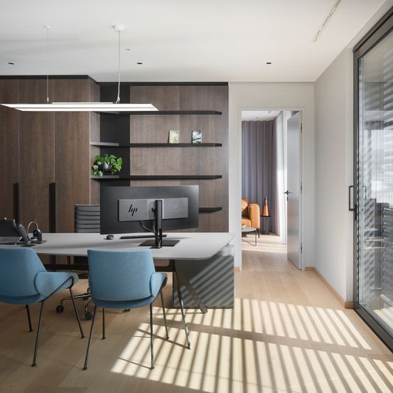 Ein leeres Büro mit einem Schreibtisch, zwei blauen Stühlen, einem Monitor, einer Topfpflanze und einer Glastür, die zu einem anderen Raum führt.