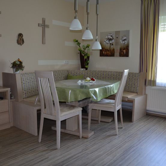 Ein Essbereich mit einem in Grün bedeckten Tisch, flankiert von zwei Stühlen. Der Raum verfügt über Holzboden, ein Fenster mit Vorhängen und eine Bank mit einem grünen Kissen.