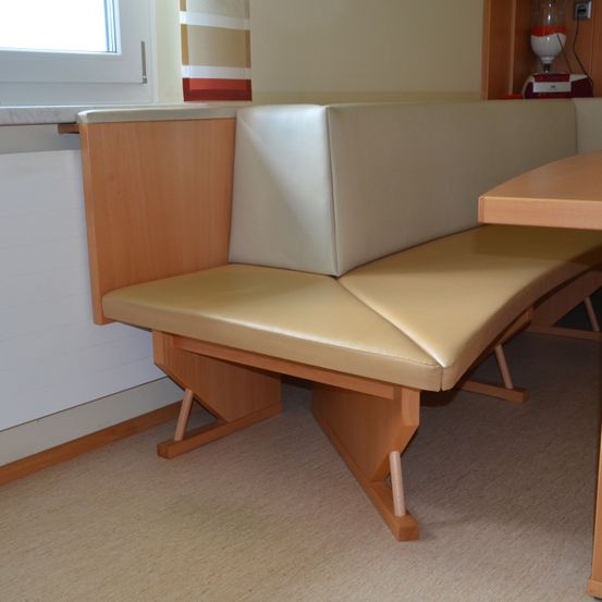 Eine beige Ledersitzbank mit Holzstützen in einer Ecke, neben einem Holztisch, in der Nähe eines Fensters mit einem gestreiften Vorhang.