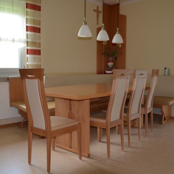 Ein Essbereich mit einem Holztisch und Stühlen, einer Bank und drei hängenden Lampen. Es gibt ein Fenster mit gestreiften Vorhängen und ein Kreuz an der Wand.