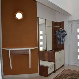 An entryway with a wooden wardrobe, a bench, a mirror, and a brown wall with a light fixture. A plaid shirt hangs on a hanger.