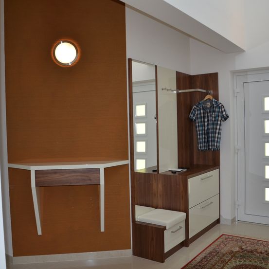 Ein Eingangsbereich mit einem hölzernen Kleiderschrank, einer Bank, einem Spiegel und einer braunen Wand mit einer Leuchte. Ein kariertes Hemd hängt an einem Kleiderbügel.