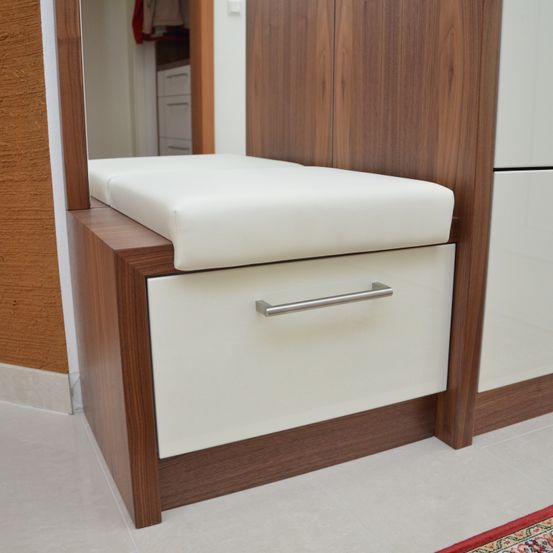 Ein hölzerner Schrank mit einem weißen Kissen und einer Schublade darunter. Der Schrank hat einen Spiegel links und eine Wand rechts.