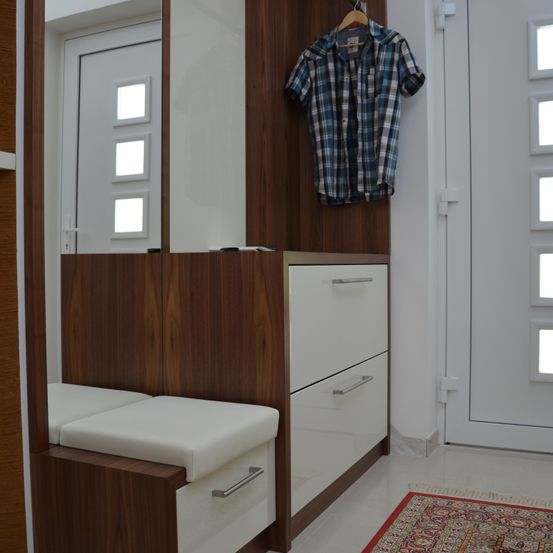 Ein moderner Eingangsbereich mit einer Holzwand, einer weißen Tür, einer Bank und drei Schubladen. Ein kariertes Hemd hängt an einem Haken.