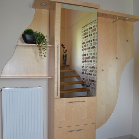 Ein hölzerner Schrank mit Spiegel zeigt eine Treppe. Links befindet sich ein weißer Heizkörper. Der Schrank hat ein Regal mit einem schwarzen Topf und einer Pflanze.