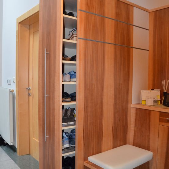 Ein Holzschrank mit Regalen ist in einem Flur zu sehen. Der Schrank ist mit Schuhen gefüllt und vor ihm steht eine weiße Bank. Links befindet sich ein Heizkörper und rechts ein Schreibtisch mit Büchern.