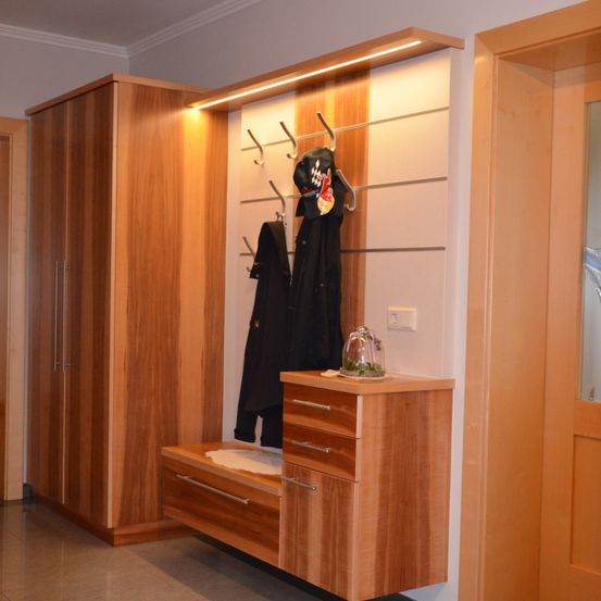 Ein Flur mit Holzschränken, einer Bank und einem Kleiderständer mit Mänteln und einem Hut. Der Raum hat eine weiße Wand und eine Holztür.