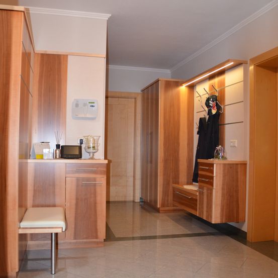 Innenansicht eines Raumes mit Holzschränken, einer Theke, einem Hocker und einem Kleiderständer. Der Raum hat einen gefliesten Boden und weiße Wände.