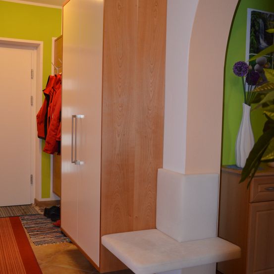 Ein Raum mit einem hölzernen Schrank und einer weißen Bank neben ihm. Ein roter Mantel hängt an einem Haken. Eine weiße Vase mit lila Blumen steht auf einem Holzschrank.