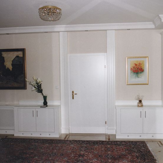 Ein Innenraum einer Wohnung mit einer weißen Tür, einer Kristallleuchte und zwei gerahmten Gemälden. Es gibt weiße Schränke mit Blumen und einem dekorativen Objekt oben. Ein Teppich bedeckt den Boden.