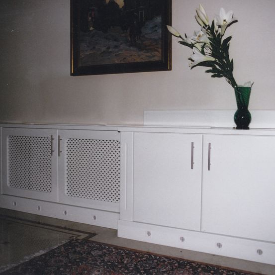 Ein Raum mit weißen Schränken und einer grünen Vase mit weißen Blumen auf einem weißen Regal über den Schränken. Über den Schränken ist ein gerahmtes Bild an der Wand befestigt.