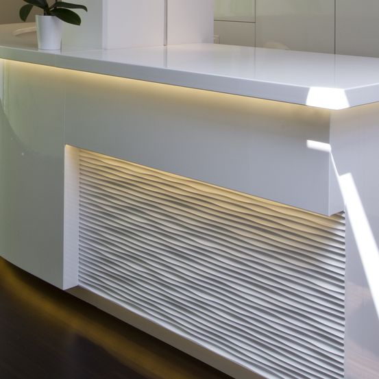 Eine weiße Rezeptionstheke mit einer Topfpflanze oben und LED-Beleuchtung entlang der Kante. Die Theke hat eine gebogene Vorderseite mit einem welligen Muster.