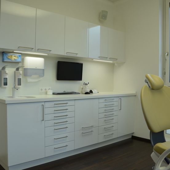 Ein modernes Zahnarztzimmer mit weißen Schränken, einem beigen Zahnarztstuhl, einem Waschbecken, einem Monitor und einem Papiertuchspender.
