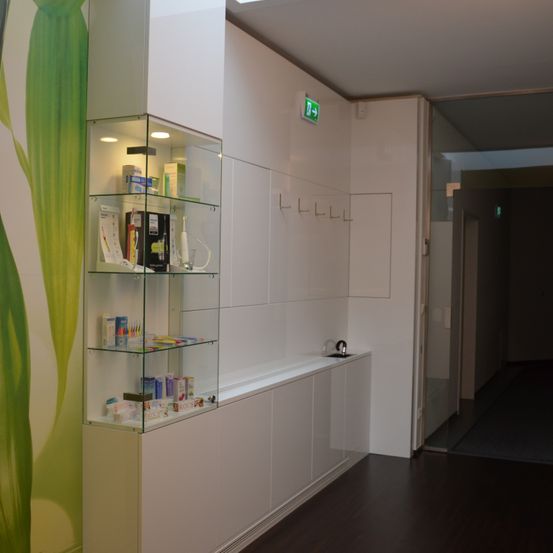 Ein Flur mit weißen Wänden und einem Glasregal mit ausgestellten Artikeln. An den Wänden befindet sich ein großes grünes Blattdesign. Es gibt eine geschlossene Tür und ein offenes Ausgangsschild.