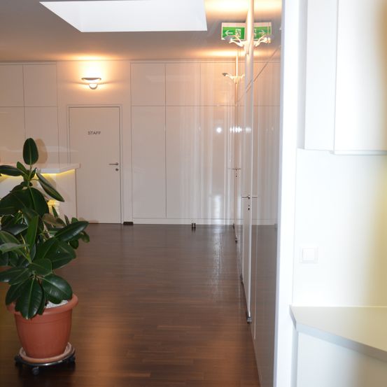 Ein leerer Flur mit weißen Wänden, einem Topf mit einer Pflanze und einer Tür mit der Aufschrift 'STAFF'. Der Boden ist aus Holz und sauber. Über dem Flur befinden sich Notausgangsschilder.