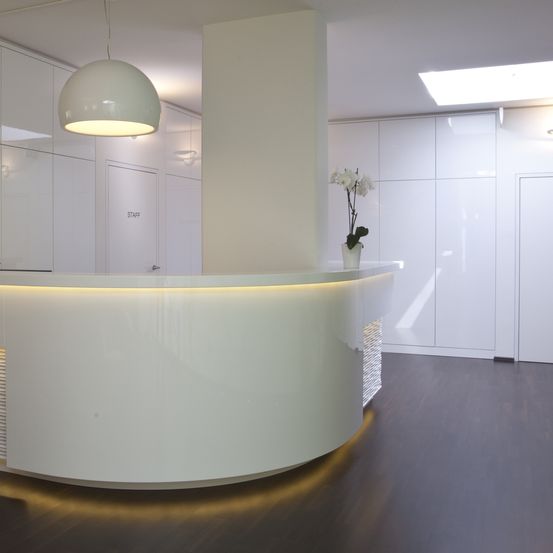 Ein Büro-Empfangsbereich mit einem modernen, gebogenen Schreibtisch. Weiße Schränke, eine große Lampe und eine Topfpflanze mit weißen Blüten schmücken den Raum. Der Boden ist aus dunklem Holz.