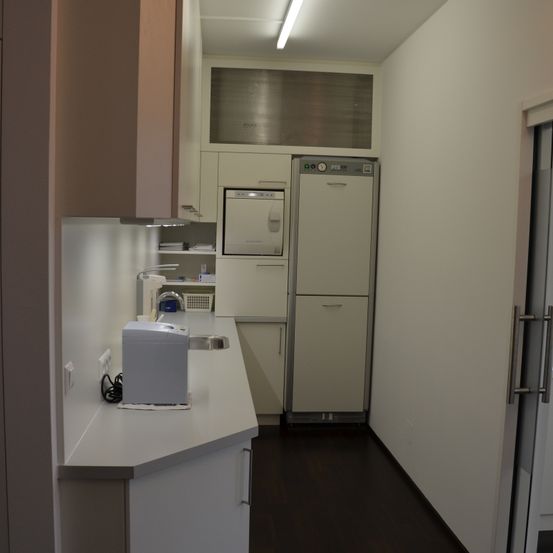 Ein sauberer, moderner Küchenbereich mit weißen Schränken, einer weißen Arbeitsplatte, einem Kühlschrank und einer Spülmaschine. Ein Waschbecken mit Wasserhahn steht auf der Arbeitsplatte. Eine Steckdose ist an der Wand montiert.