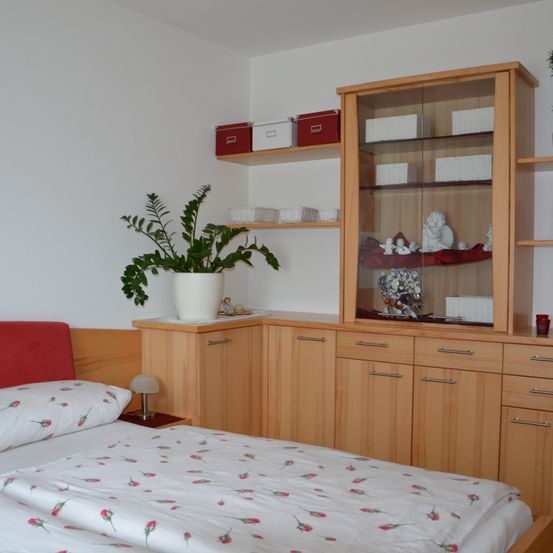 Ein Schlafzimmer mit einem Bett, das mit einer Blumendecke bedeckt ist, einem Nachttisch mit einer Lampe und einem Holzschrank mit Glastür. Ein Topf mit Pflanzen steht auf dem Regal über dem Bett.