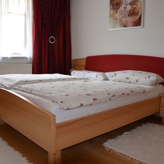 Ein Schlafzimmer mit zwei Holzbetten, roten Kopfteilen, weißen Laken und Blumenkissen. Ein weißer Heizkörper und rote Vorhänge schmücken das Fenster. Ein gerahmtes Gemälde hängt an der Wand.