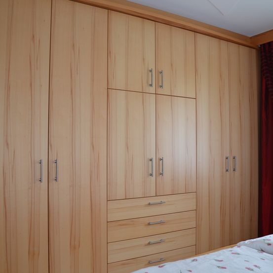 Ein Schlafzimmer mit einem großen hölzernen Kleiderschrank, ausgestattet mit mehreren Türen und Schubladen, beleuchtet durch natürliches Licht von oben. Das Bett ist mit einem weißen, blumengemusterten Laken bedeckt.
