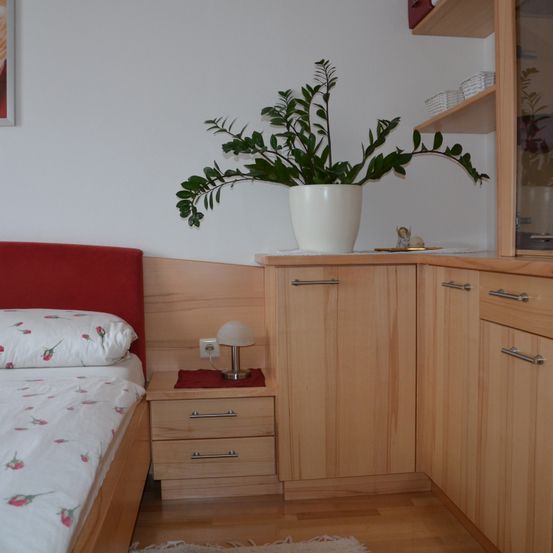 Ein Schlafzimmer mit Holzmöbeln, einem Bett mit bedruckter Bettwäsche, einem Nachttisch mit Lampe und einer weißen Topfpflanze auf einem Schrank.