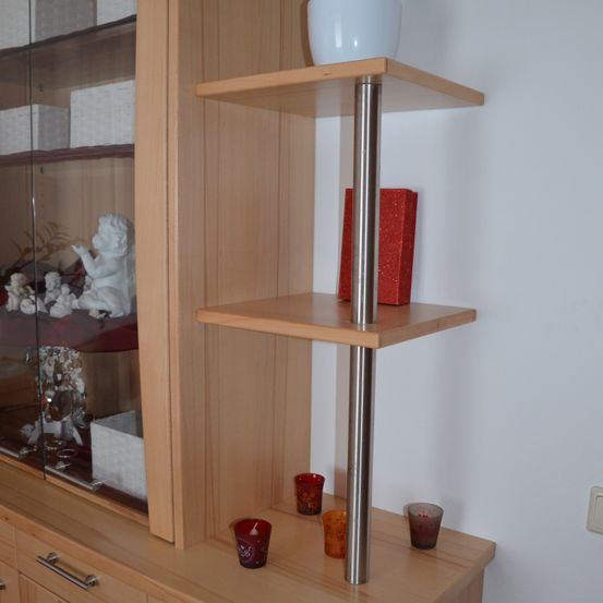 Ein hölzerner Schrank mit einem Metallpfosten steht an einer weißen Wand und zeigt Dekorationsartikel auf zwei Regalen. Das obere Regal enthält eine weiße Vase und eine rote Schachtel, während das untere Regal drei rote Kerzenhalter aufweist.