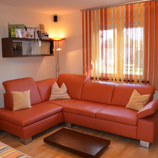 Ein modernes Wohnzimmer mit einem orangefarbenen Ledersofa, Kissen, einem Holztisch und einer Stehlampe. Ein Regal mit Büchern und einem Lautsprecher befindet sich an der Wand. Das Fenster hat orangefarbene Vorhänge.