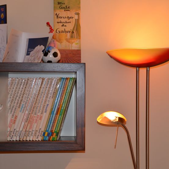 Ein Bücherregal mit Büchern und einer Lampe, mit einem Poster, das 'Alles Gute zum Geburtstag' liest. Die Lampe befindet sich auf der rechten Seite und wirft ein orangefarbenes Licht an die Wand.