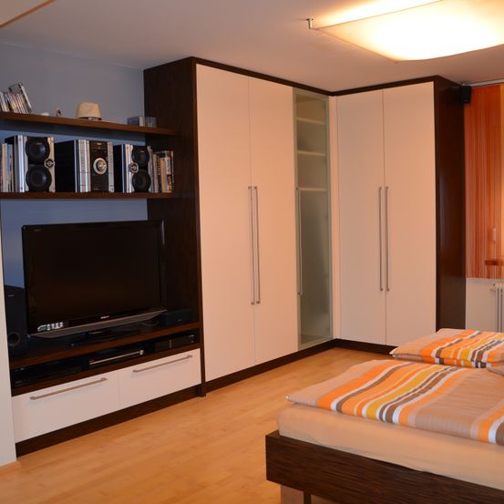 Ein modernes Schlafzimmer mit einem Flachbildfernseher auf einem Schrank, zwei Betten mit farbenfrohen gestreiften Bettwäschen, Einbauschränken, einer Deckenlampe und einem Holzboden.