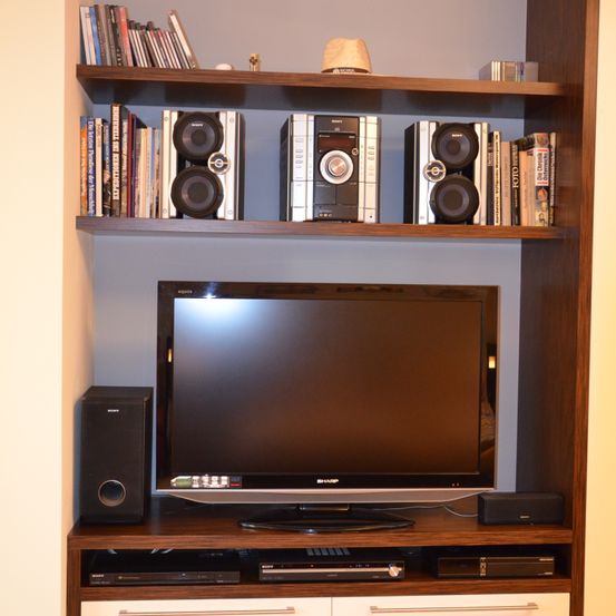 Ein Unterhaltungszentrum aus Holz mit einem Flachbildfernseher und Lautsprechern. Drei Sony-Lautsprecher befinden sich im oberen Regal mit Büchern und einer Mütze. Ein schwarzer Lautsprecher ist links unten am Fernseher.