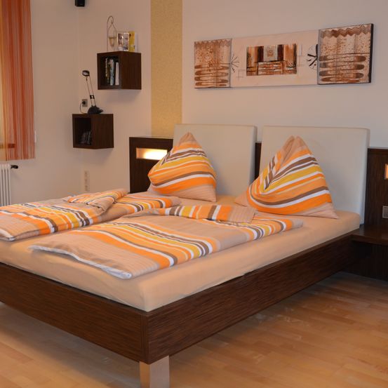 Ein Schlafzimmer mit einem Bett, zwei Kissen und einem weißen Kopfteil. Drei abstrakte Gemälde hängen an der Wand. Eine Lampe steht auf einem Regal.