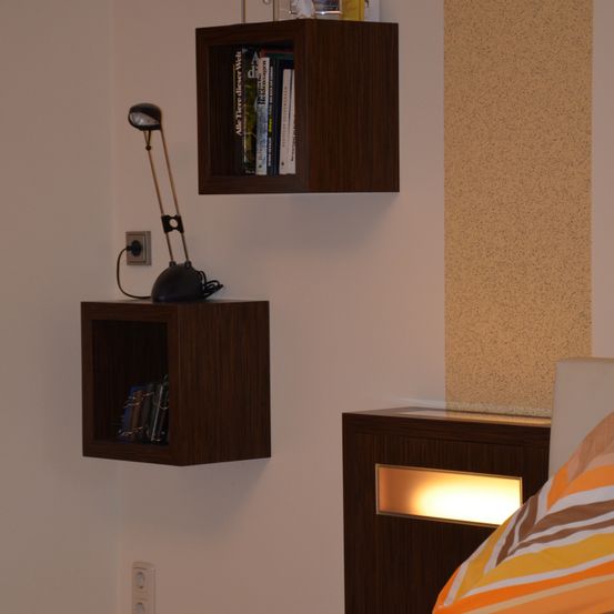 Ein Schlafzimmer mit zwei schwebenden Regalen, eines mit Büchern und einer Lampe, das andere mit einer beleuchteten Lampe. Die Wand ist weiß und ein Bett ist teilweise zu sehen.