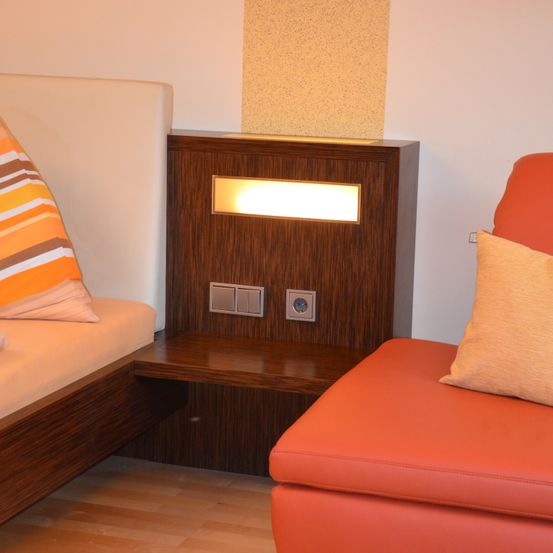 Ein kleiner Raum mit zwei Betten und einem orangefarbenen Sofa. Jedes Bett hat ein Kissen. Die Wand hat ein integriertes Licht.