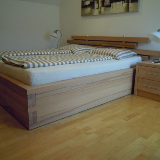 Ein Schlafzimmer mit einem Holzbett, zwei Matratzen und Kissen. Ein Nachttisch mit einer Lampe befindet sich auf der rechten Seite. Ein gerahmtes Bild ist an der Wand.