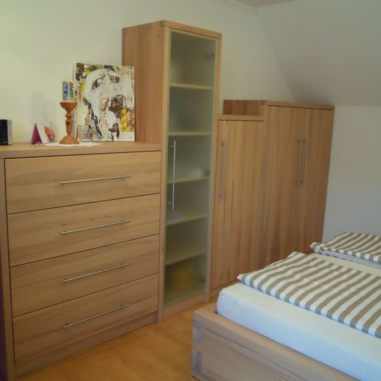 Ein Schlafzimmer mit Holzmöbeln umfasst eine Kommode, einen Schrank, eine Glasvitrine und ein Bett mit gestreiften Laken. Eine Kerze steht auf der Kommode.