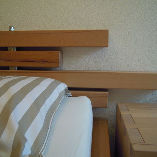 Ein Bett mit hölzerne Kopfteil und einem gestreiften Kissen an einer weißen Wand. Ein hölzernes Nachttisch mit Schubladen ist in der Nähe.
