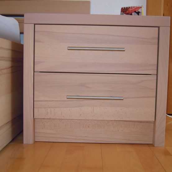 Ein hölzerner Nachttisch mit zwei Schubladen, auf einem Holzboden neben einem Bett platziert.