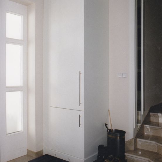 Ein Innenansicht eines Flurs mit weißen Wänden, einem hohen weißen Schrank, einem Fenster und einer Treppe. Ein Besen und Kehrschaufel stehen neben dem Schrank.