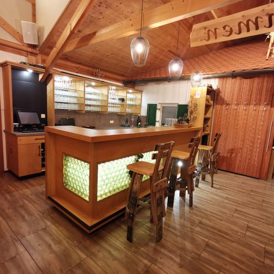 Ein Innenraumblick einer rustikalen Bar mit Holzarbeitsplatten, Regalen und hängenden Lampen. Es gibt Stühle und Hocker um die Bar und eine Glasvitrine.