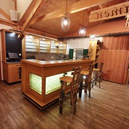 An interior view of a rustic bar with wooden countertops, shelves, and hanging lamps. There are chairs and stools around the bar, and a glass display case.