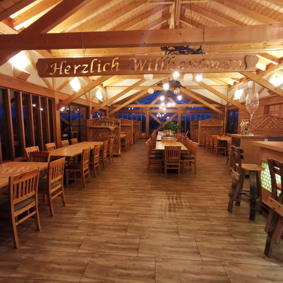 Ein Restaurant mit Holzboden und Decke, Holztischen und Stühlen. Ein Schild mit Herzlich Willkommen ist an der Decke angebracht.