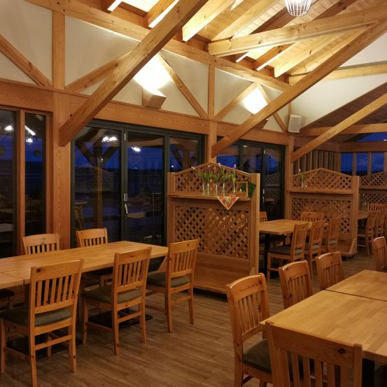 Ein leerer Innenbereich zum Essen mit Holztischen und -stühlen, Holzwänden und Glastüren. Es gibt Holzkabinen und eine Holzabtrennung mit Blumen oben.
