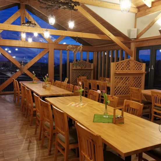 Ein Essbereich mit Holztischen und Stühlen, langen Fenstern und dekorativen Elementen. Blumen stehen in Vasen auf den Tischen und Lampen hängen von der Decke.